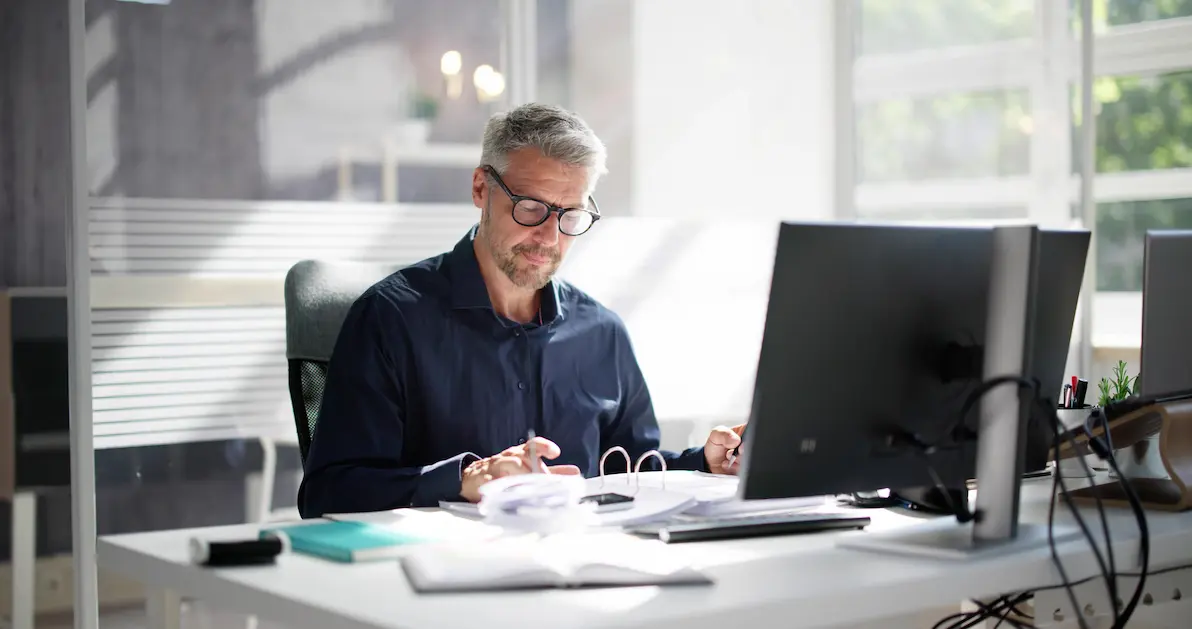 A man with glasses working at a desk with a laptop and papers.