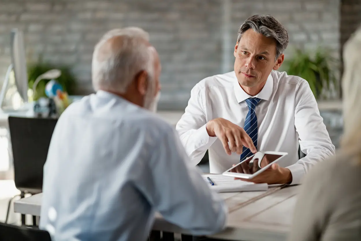 Two men in a professional meeting, one holding a tablet and gesturing, seated at a table.